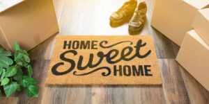 Home Sweet Home" doormat surrounded by unpacked boxes, a plant, and a pair of shoes on a wooden floor.